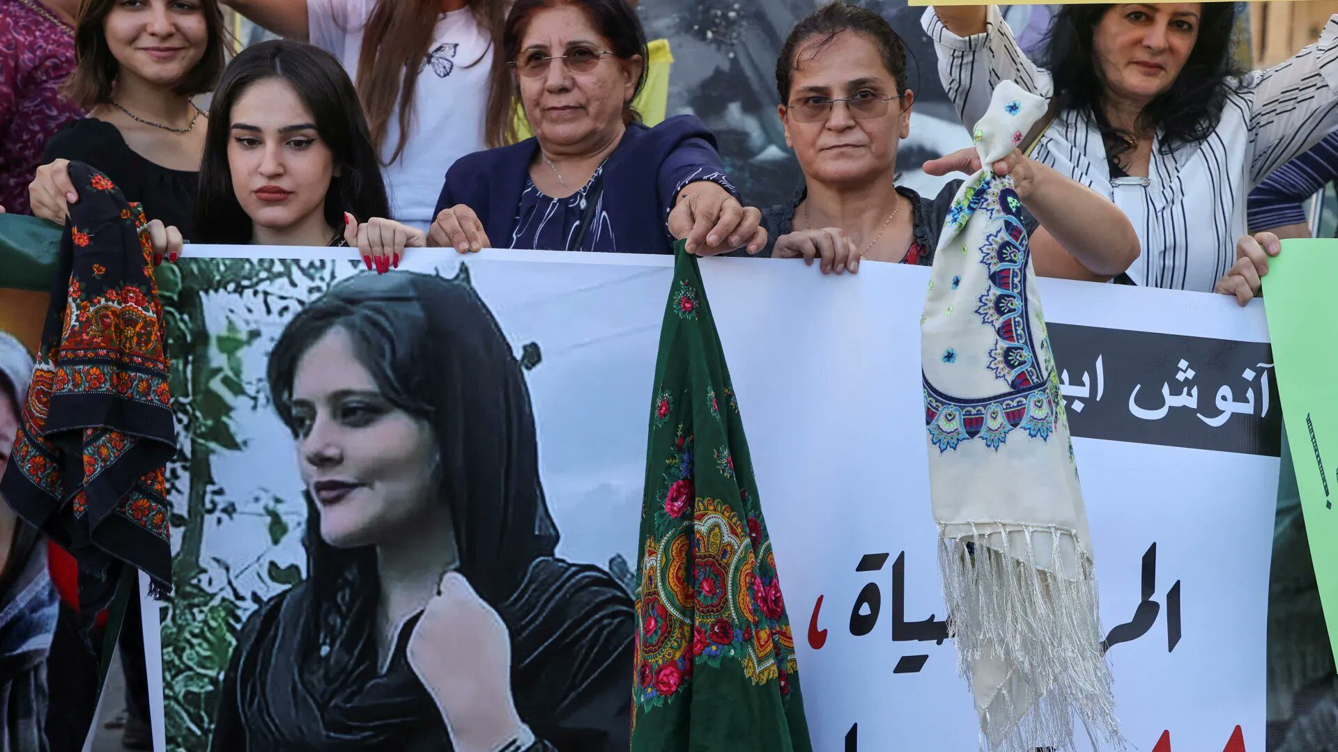 Women hold a portrait of Mahsa (Jina) Amini and headscarves during Woman, Life, Freedom protests.