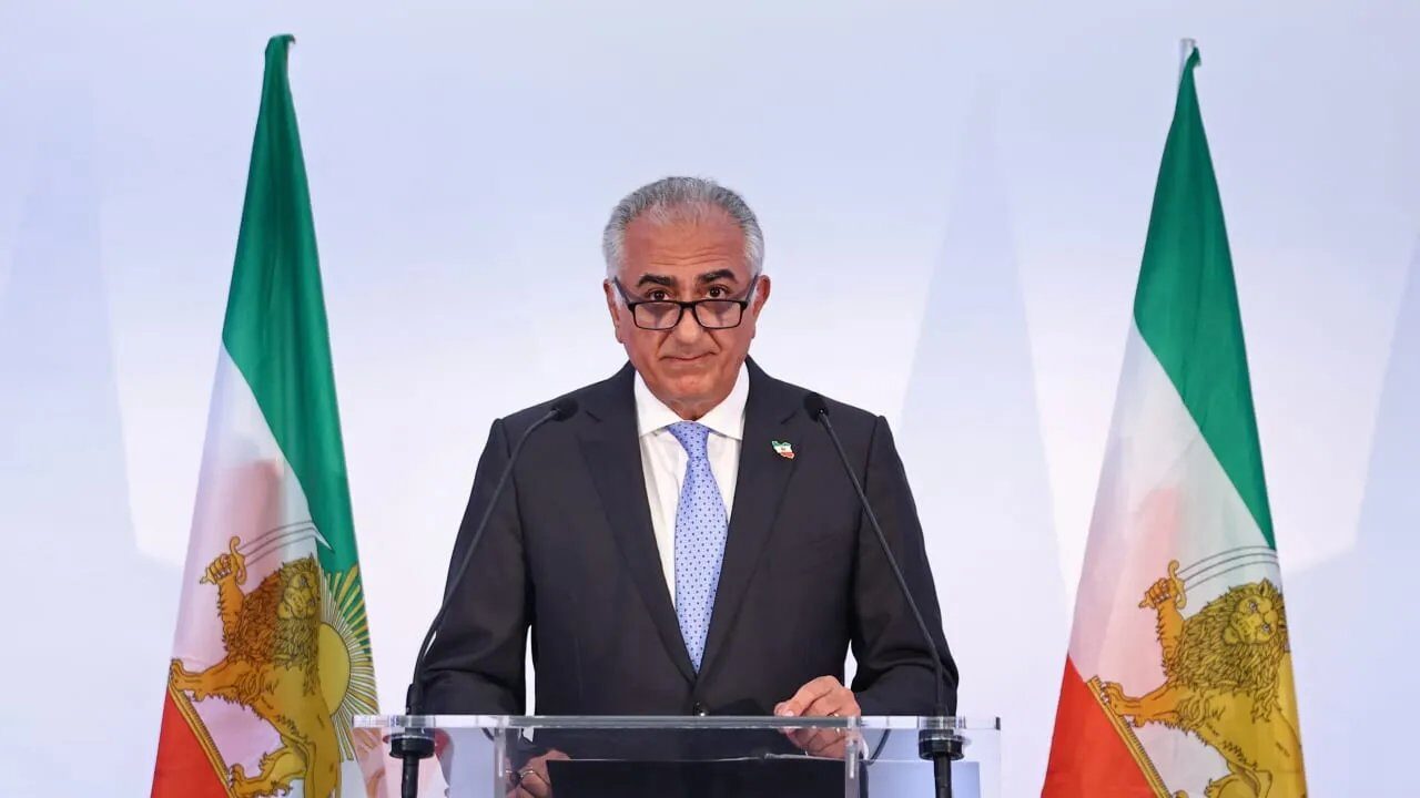 Reza Pahlavi at a podium flanked by Lion and Sun flags while outlining his vision for a transitional system.